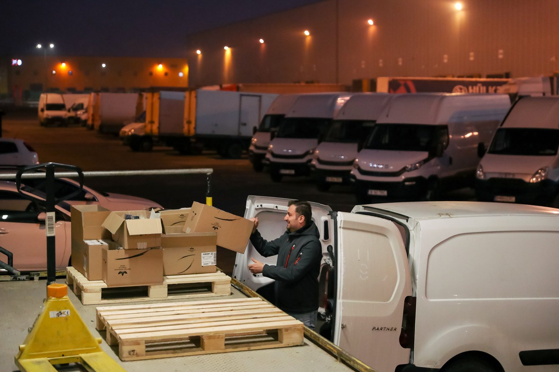 Reportaj de noapte într-un centru logistic: un om încarcă o dubă lângă paleți, cu flota de vehicule în fundal